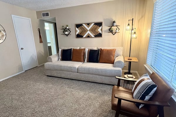 Bright living room with beige sofa, geometric wall art, pendant lights, and wood accent chair near shuttered windows