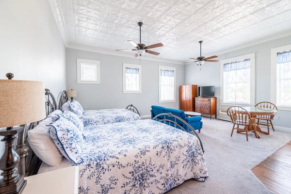 Spacious bedroom with two beds, blue patterned quilts, ceiling fans, wooden furniture, and sitting area with blue armchair