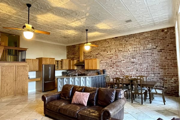 Open-plan living room with brown leather sofa, exposed brick wall, wood kitchen cabinets, and decorative tin ceiling.